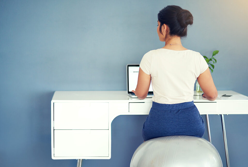 View from behind of woman sitting on balance ball chair at a desk working on a laptop.