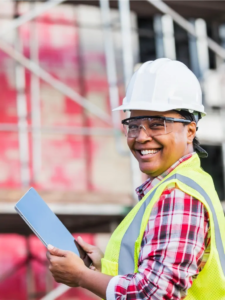 Smiling maintenance worker wearing yellow safety vest and hard hat holding tablet on site.