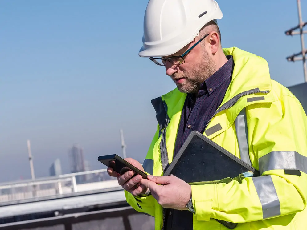 Inspector on site wearing yellow safety jacket and hard hat holding tablet under arm and using mobile phone in hands.