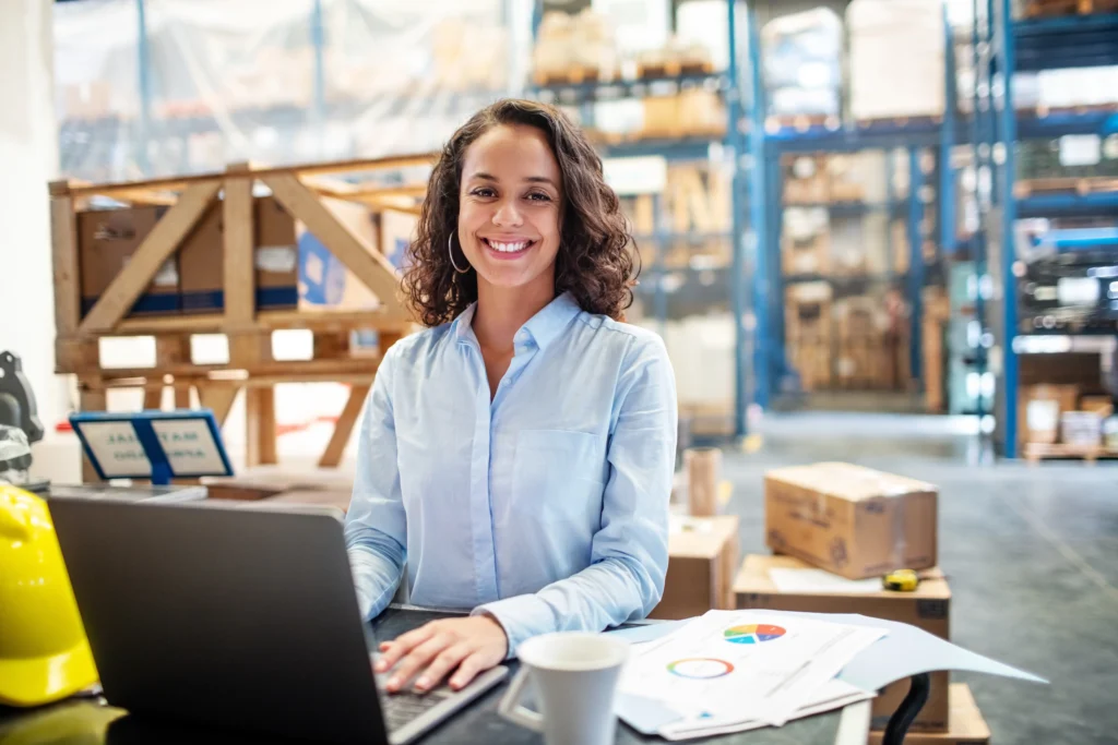 Smiling woman in large warehouse using laptop.