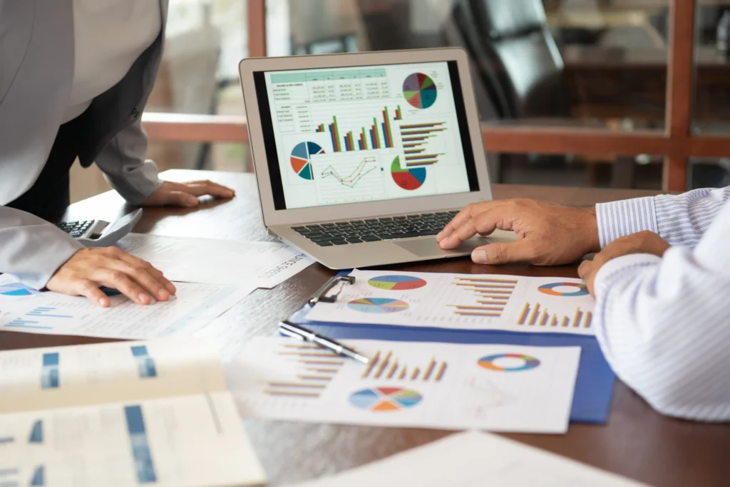 Hands of two colleagues leaning on an office desk reviewing charts and graphs on a laptop.