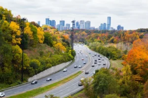 Cars driving on highway with beautiful Autumn coloured trees on either side and city skyline in the horizon.