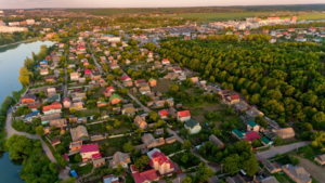 Aerial view of residential area with green wooded area on the right side and small body of water on the left side on a sunny day.