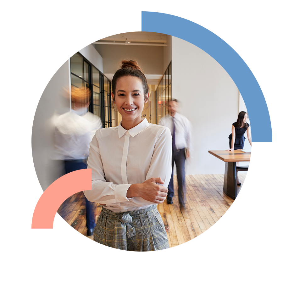 Confident, professional woman standing in modern office smiling while colleagues have a discussion in the background.