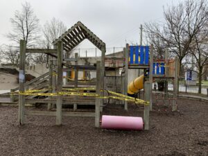 Wooden playground equipment blocked with yellow caution tape on a rainy day.