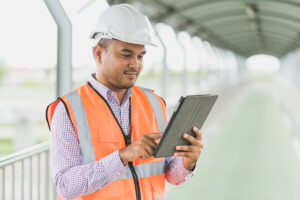 Inspector on site wearing orange safety vest and white hard hat using digital tablet on site.