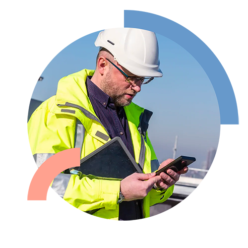 Inspector on site wearing yellow safety jacket and hard hat holding tablet under arm and using mobile phone in hands.