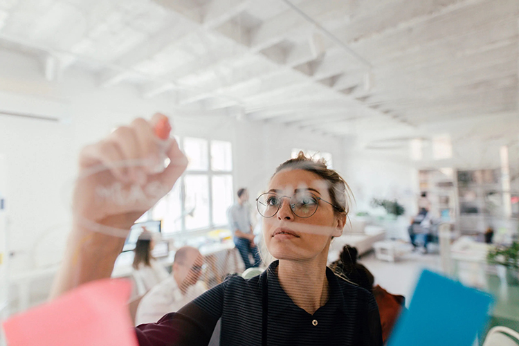 Looking through an interior office window at woman writing notes on the glass surface with a marker.