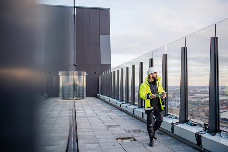 Inspector wearing yellow safety jacket, golding digital tablet while walking along high rise balcony with glass railings.