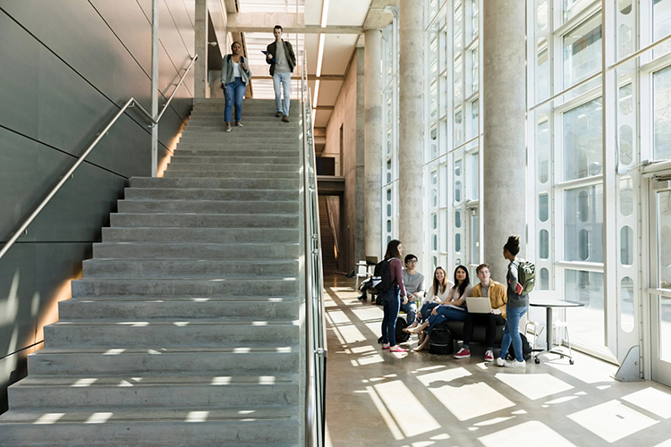 Group of students taking a break in the hall way of a campus building while two other students walk down a set of stairs.