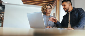 Two colleagues in an office having a discussion while looking at a laptop.