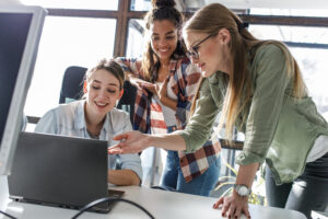 A senior programmer guides a beginner at a laptop while a diverse team works nearby.