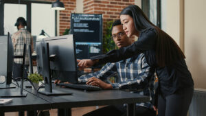 Two colleagues collaborating at a computer workstation in a modern office, discussing maintenance management software displayed on the monitor.