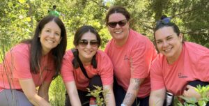 Four employees planting a tree together during an employee volunteer program in a green outdoor setting.